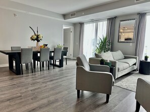 A modern living room with a grey sofa, a dining table with chairs, and a large window with curtains. - Next Level Living in Niagara! (Niagara Falls)