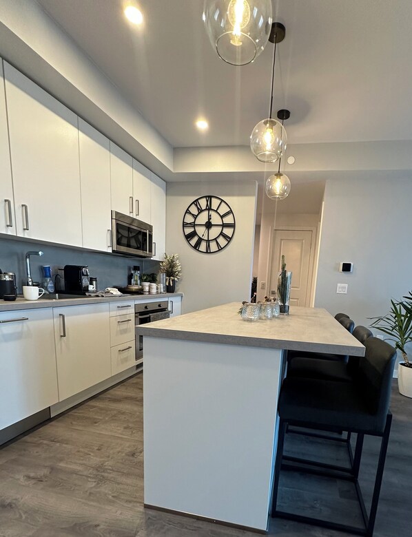Una cocina moderna con armarios blancos, una isla central, iluminación colgante y un reloj de pared grande.