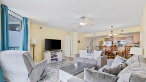 A living room with a sofa, coffee table, and TV. There is a kitchen in the background. - Stay Elevated – North Myrtle Beach 3BR Penthouse at Malibu Pointe Resort (North Myrtle Beach)