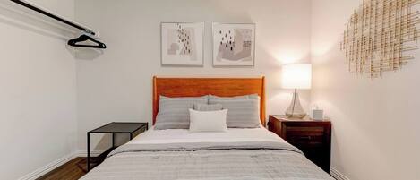 A hotel room with a bed, a nightstand, a wall-mounted lamp, and two framed artworks.