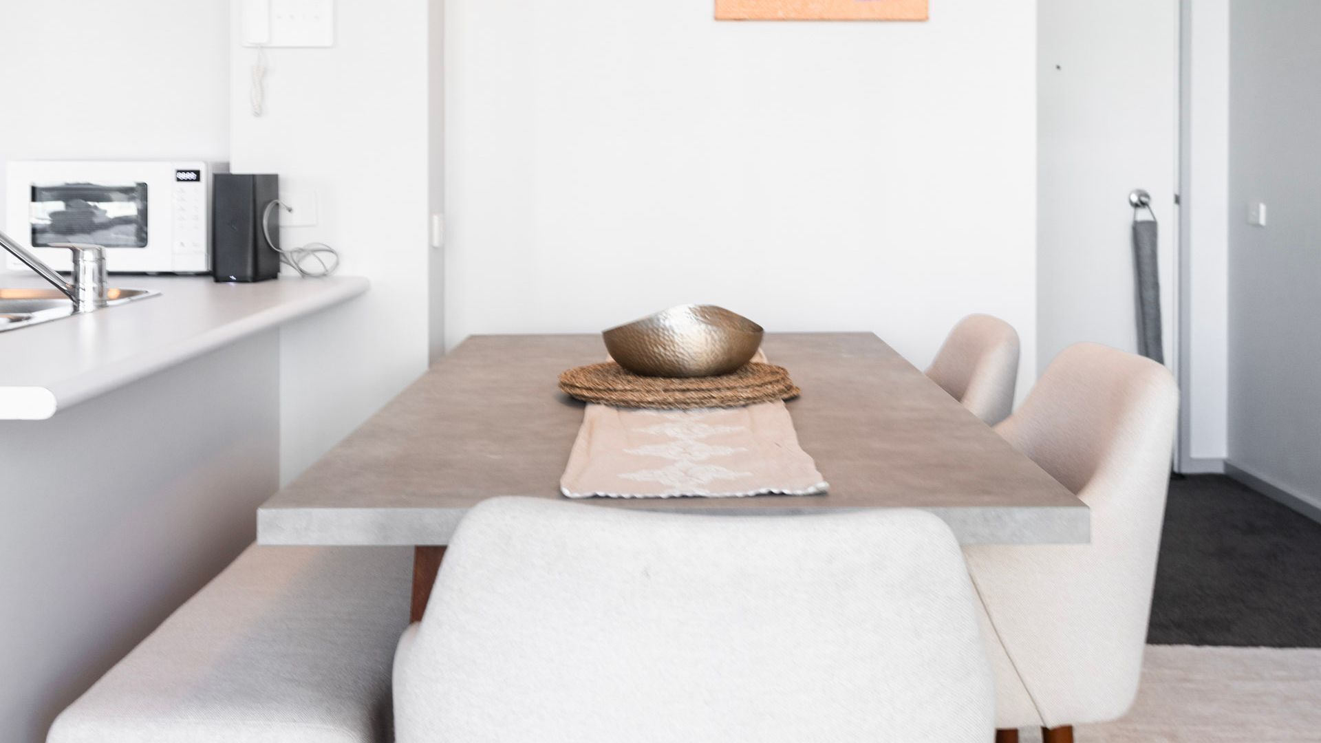 Un comedor moderno con una mesa rectangular, sillas blancas y una zona de cocina con microondas y hervidor.