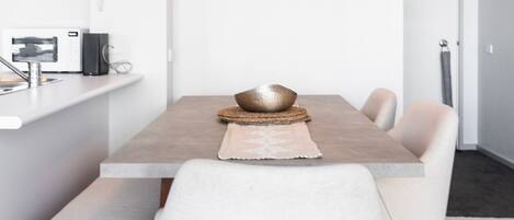Un comedor moderno con una mesa rectangular, sillas blancas y una zona de cocina con microondas y hervidor.