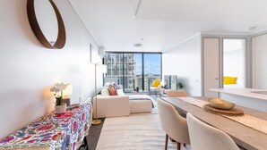 A modern apartment with a dining area, a sofa, a small table with a colorful tablecloth, and a large window offering a city view.
