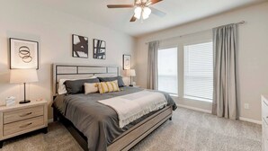 Une chambre à coucher avec un lit, des tables de chevet, un ventilateur de plafond et de grandes fenêtres avec des rideaux.