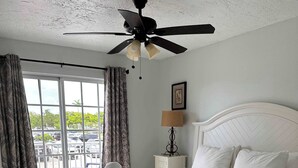 Ein Hotelzimmer mit Bett, Deckenventilator, Fenster mit Vorhängen, Nachttisch mit Lampe und Blick auf einen Parkplatz.