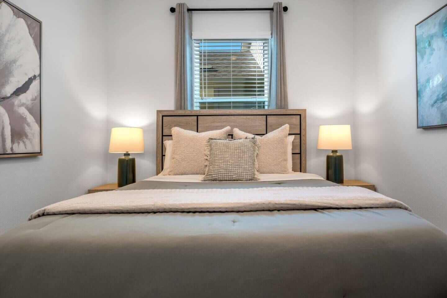 A neatly made bed with a headboard, two bedside lamps, and a window with blinds.