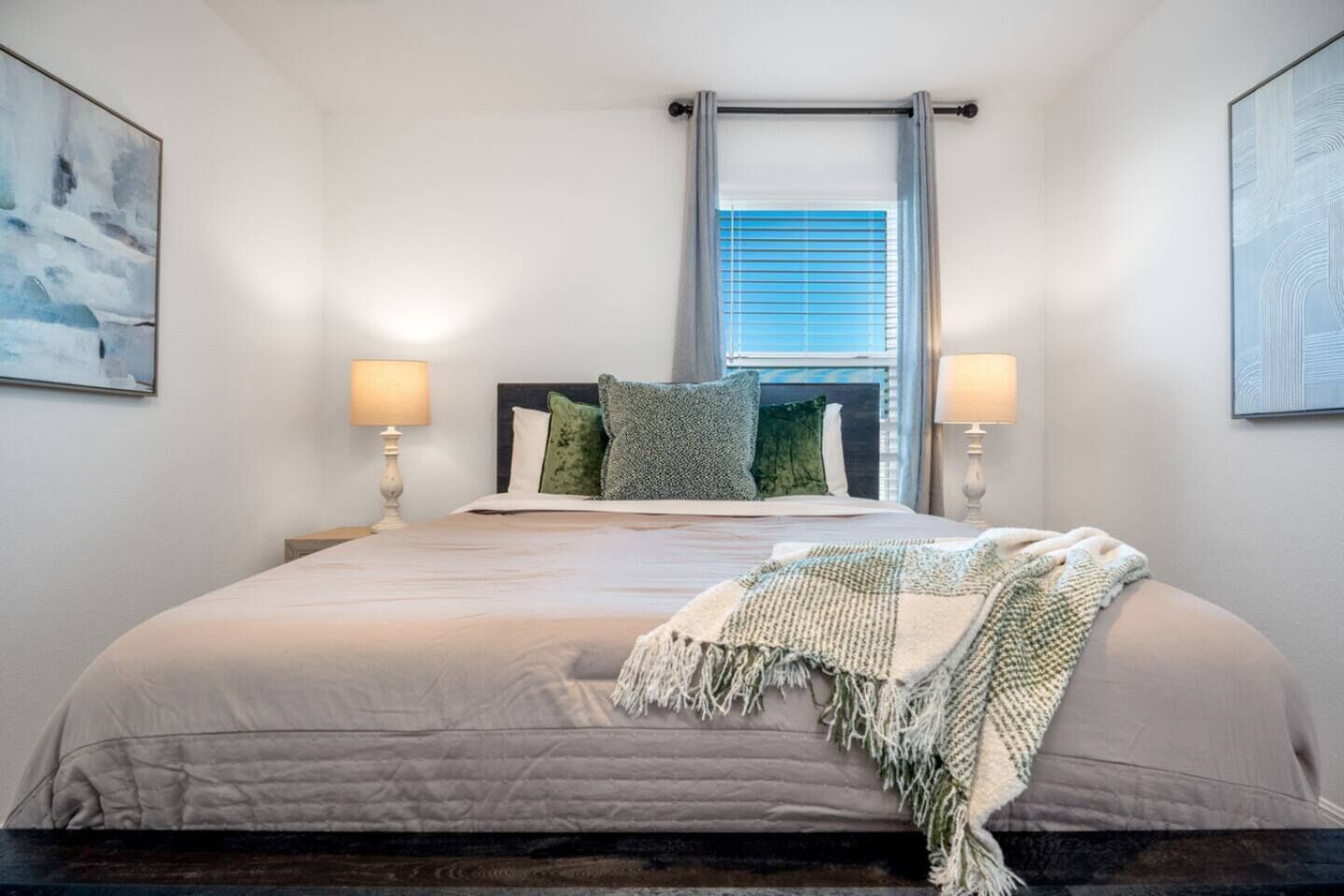 Een slaapkamer met een groot bed, twee bedlampen, een raam met zonwering en kunst aan de muren.
