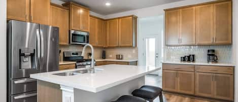 Une cuisine moderne avec des meubles en bois, des appareils en acier inoxydable, un grand îlot central avec des tabourets et un pare-feu en carreaux.