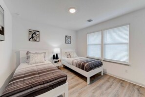 A bedroom with two single beds, a nightstand with a lamp, and two windows with blinds. - 4BR Corporate Stay in Hutto (Hutto)