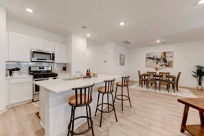 Una cocina moderna con armarios blancos, una isla central con taburetes, un comedor con mesa y sillas de madera, y un cuadro en la pared.