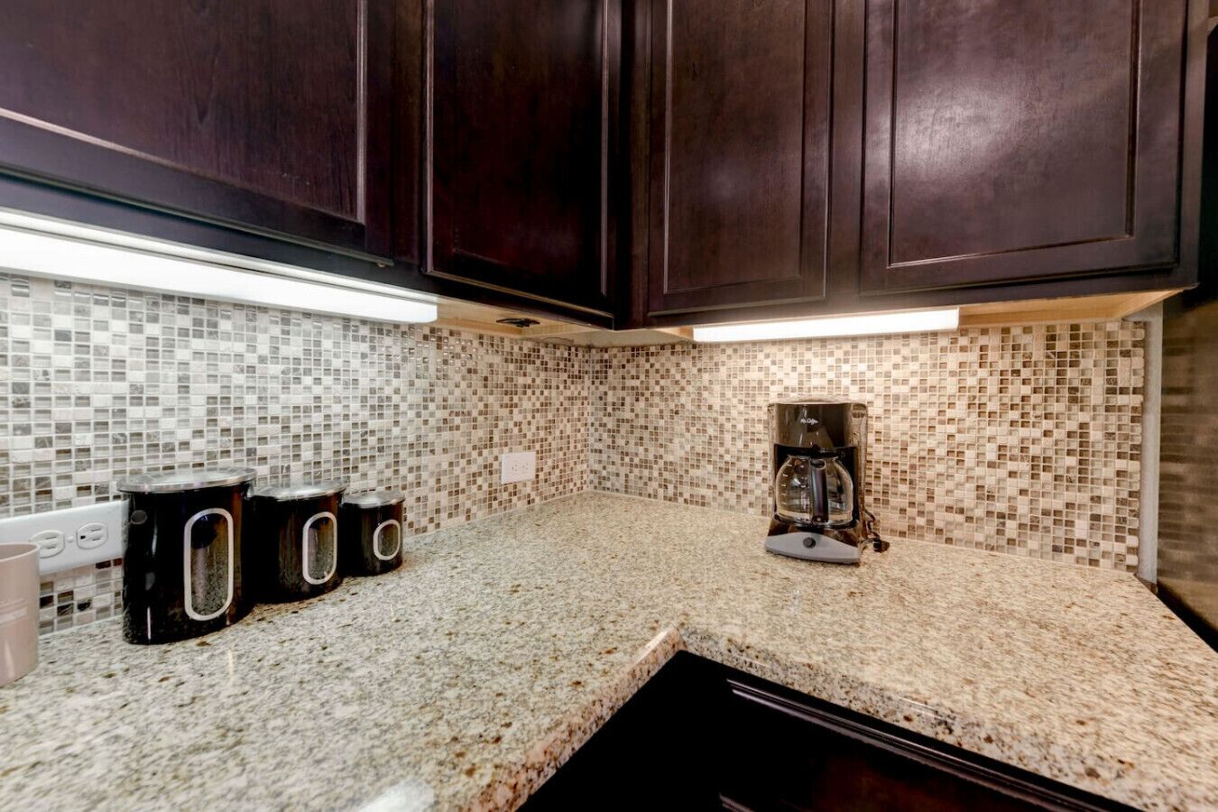 Una cocina con armarios de madera oscura, salpicadero de azulejos y encimera de granito. Hay tres cubos de basura negros y una cafetera sobre la encimera.