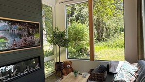 A modern living room with a fireplace, large windows, and a view of greenery. - Private Get Away TINY HOUSE with SAUNA (Stanwood)