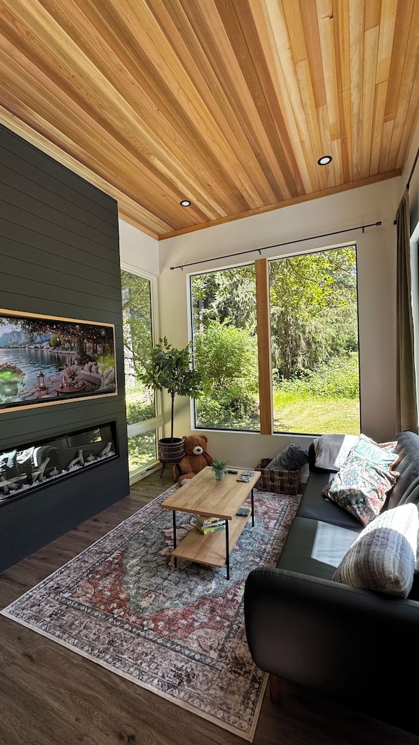 A modern living room with a fireplace, large windows, and a view of greenery.
