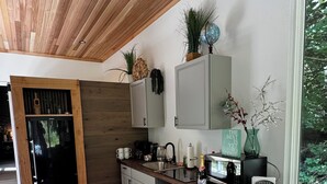 A modern kitchen with a wooden ceiling, a built-in island with bar stools, and a view of greenery outside. - Private Get Away TINY HOUSE with SAUNA (Stanwood)