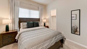 A bedroom with a bed, two bedside tables, a nightstand, a window with blinds, a door, and a mirror on the wall.