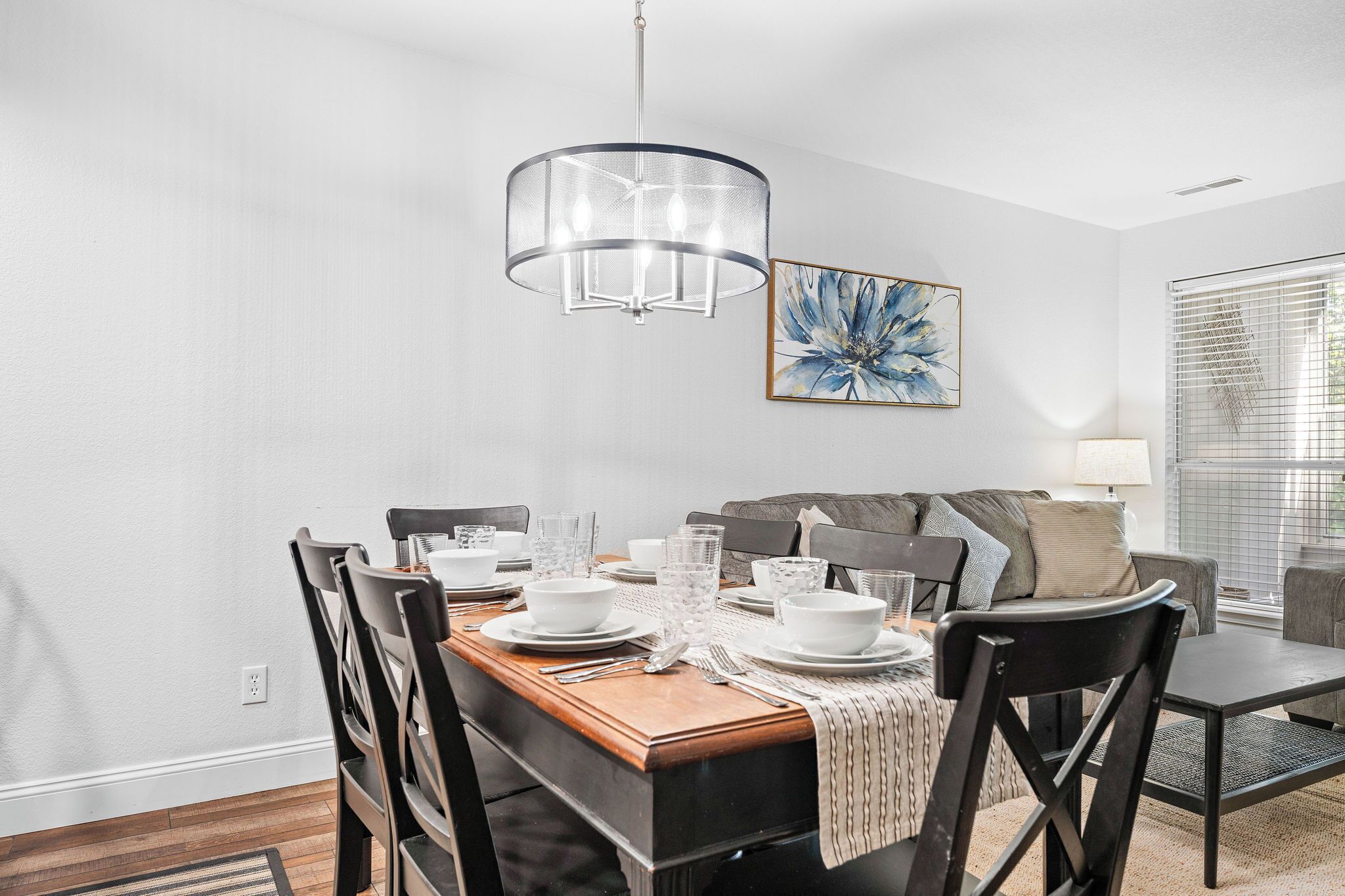 A dining area with a wooden table set for four, a modern chandelier, and a large abstract painting on the wall.