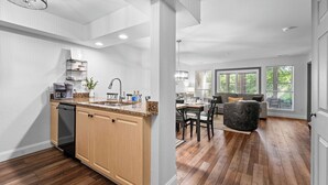 A modern kitchen with wooden cabinets, a dishwasher, and granite countertops. There is a dining area with a table and chairs, and a living room with a sofa and a large window. - Peaceful Golf Course View + Pool + Sunroom (Branson)