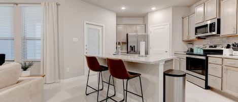 A modern kitchen with white cabinetry, a central island with bar stools, a microwave, stove, and a trash can.