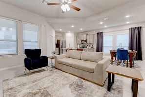 A modern living room with a sofa, a blue armchair, a dining table with chairs, and a kitchen area in the background. - Luxurious 4-Bedroom House (Temple)