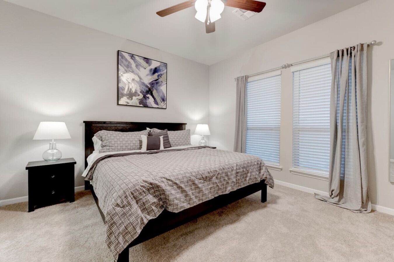 Une chambre à coucher avec un lit, deux tables de chevet avec des lampes, un ventilateur de plafond, une fenêtre avec des rideaux et un tableau au mur.