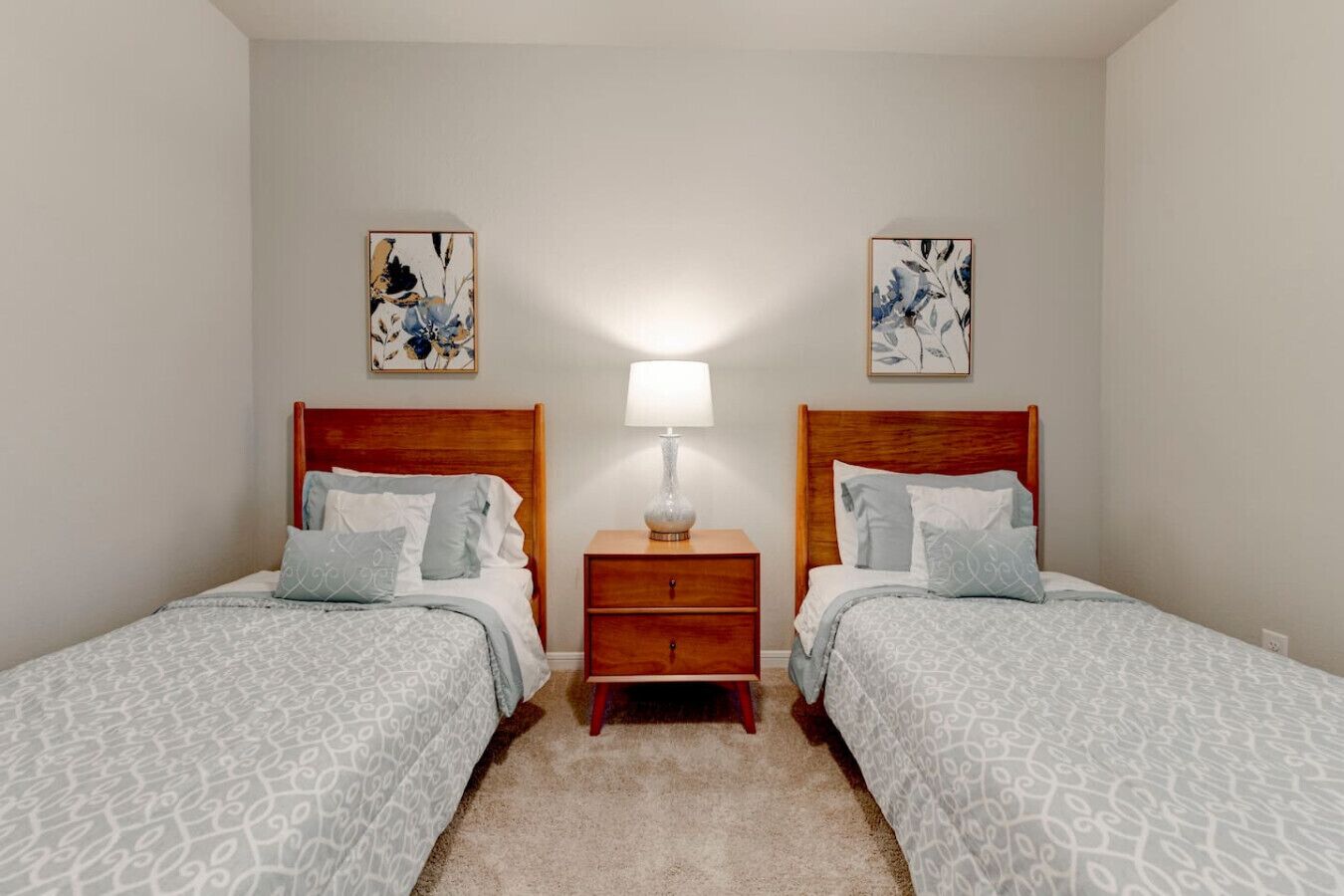 Un dormitorio con dos camas individuales, una mesita de noche con lámpara y dos cuadros colgados en la pared.