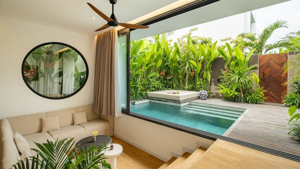 A modern living room with a large window, a ceiling fan, a round mirror, and a small round table with a plant. - Club Suma Villa Bingin (Pecatu)