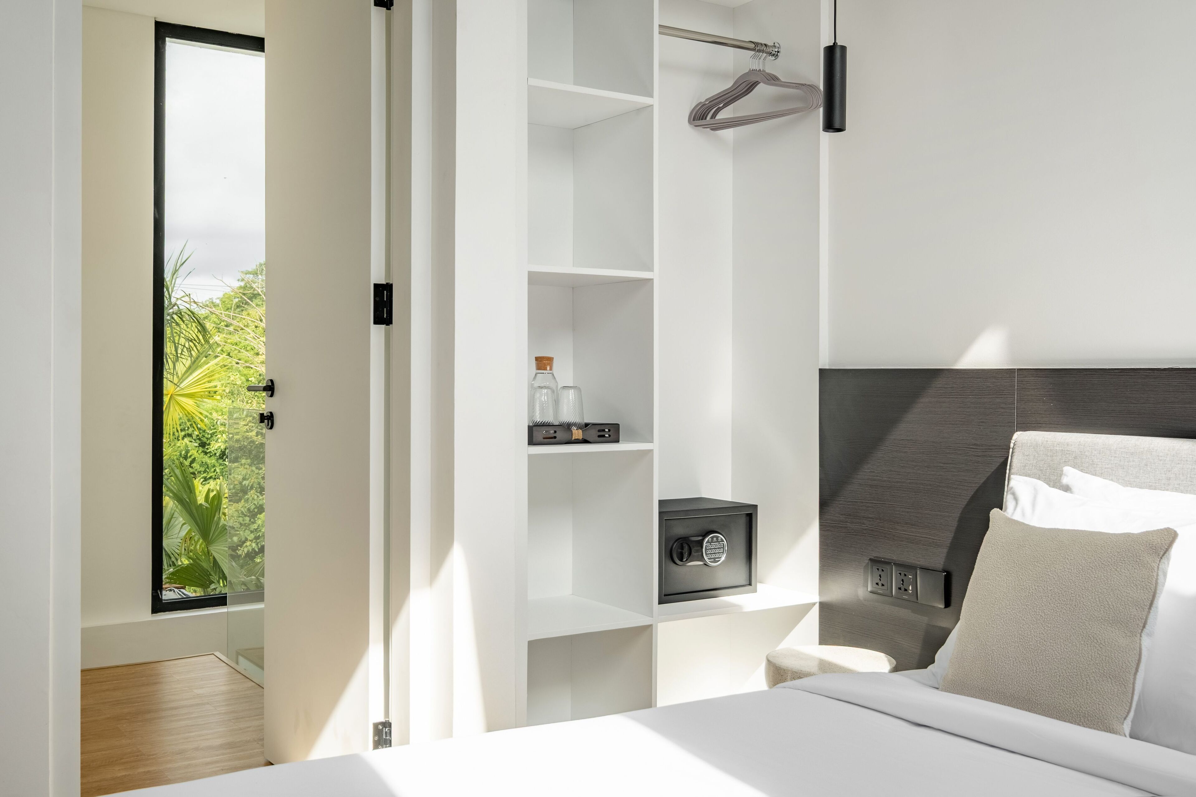 A modern bedroom with a bed, a safe, a hanging rack, and a view of greenery outside.