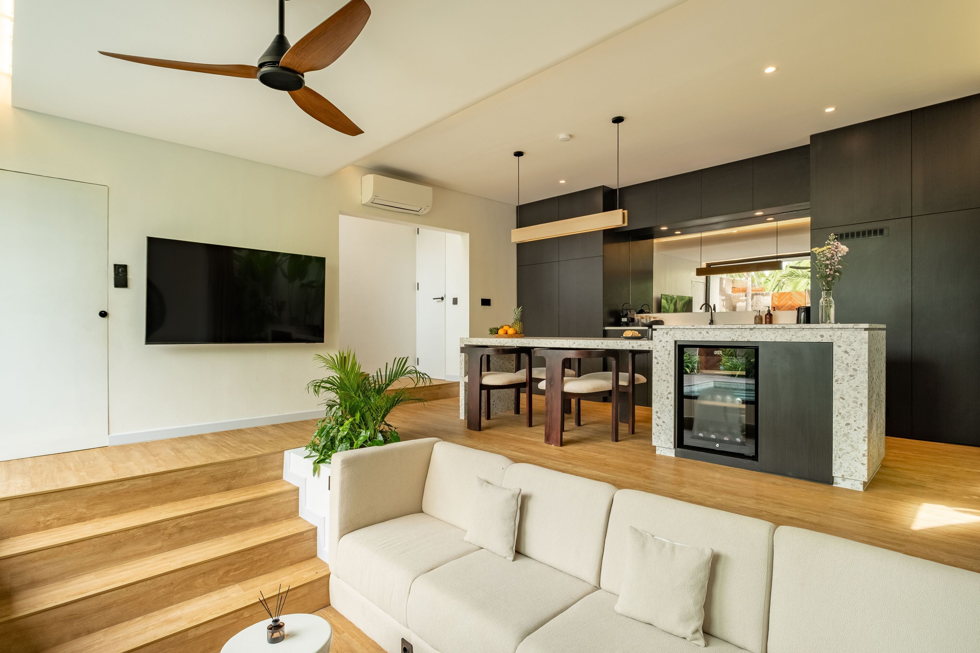 A modern living room with a sofa, a dining area with chairs, a kitchen with a countertop, and a ceiling fan.
