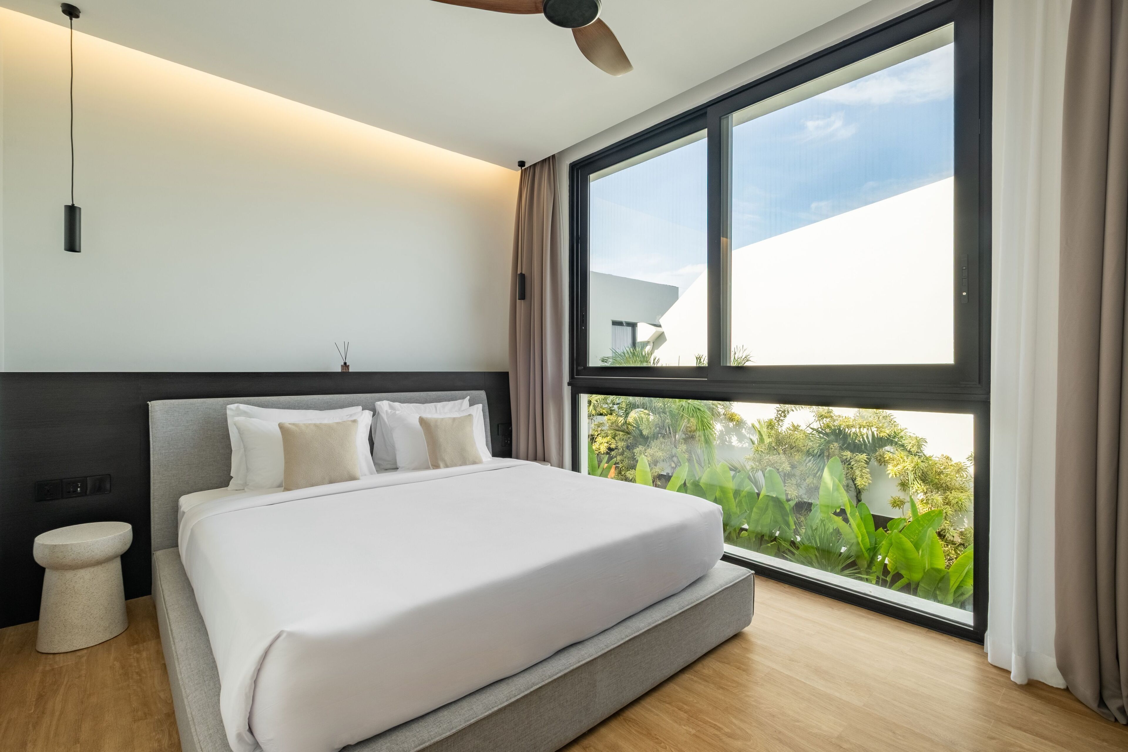 A modern bedroom with a large bed, a ceiling fan, and a window with a view of greenery.