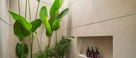 A bathroom with a shower, a shelf with toiletries, and large green plants.
