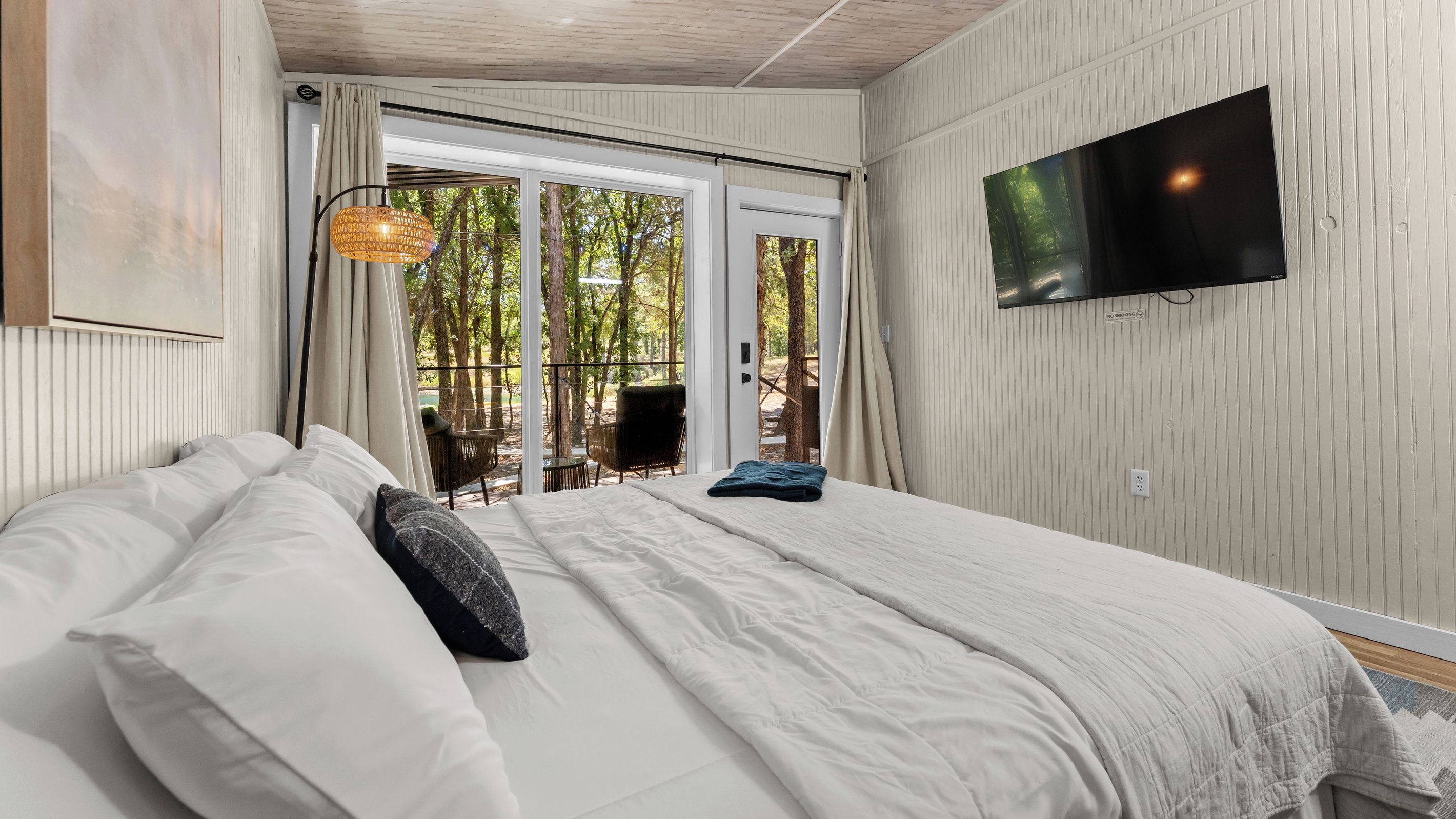A bedroom with a large bed, a TV mounted on the wall, and a view of the outdoors through a sliding door.