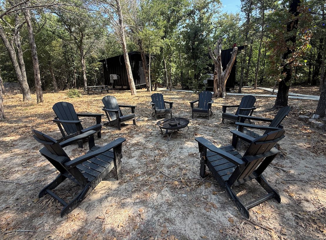 En gruppe Adirondack-stole arrangeret i en rundkreds omkring et bålsted i et skovområde med en rustik hytte i baggrunden.