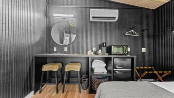 A modern room with a wooden wall, a round mirror, a small kitchenette, two wicker stools, a bed, and a trash bin.