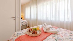 A hotel room with a bed, a breakfast tray with croissants, fruit, and orange juice, and a neatly made bed with towels and a folded robe. - Stylish & Spacious Apartment by HostWise (Porto)