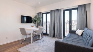 Ein modernes Wohnzimmer mit grauem Sofa, einem Esstisch mit zwei Stühlen, einem flachbildfernseher an der Wand und großen Fenstern mit grauen Vorhängen.