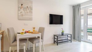 Un salon moderne comprenant une table à manger, des chaises, un téléviseur à écran plat, une petite table d’appoint avec une plante, et une vue sur le balcon extérieur.