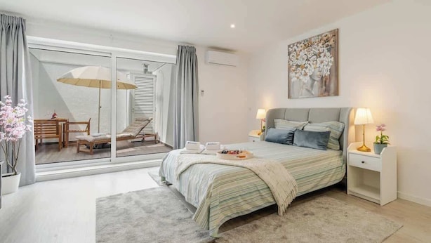 Une chambre à coucher comprenant un lit, des tables de chevet, une fenêtre avec des rideaux, un tableau et une vue sur un balcon aménagé avec du mobilier.