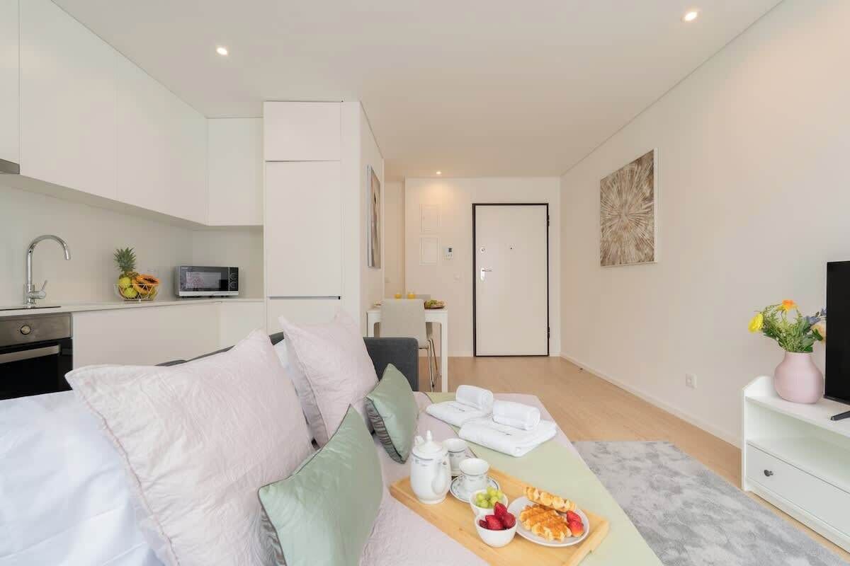 Una habitación moderna y bien iluminada, con cama, una kitchenette pequeña, un área de comedor con una bandeja de fruta y un televisor sobre un soporte blanco.
