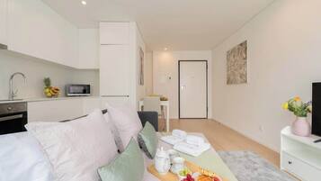 Una habitación moderna y bien iluminada, con cama, una kitchenette pequeña, un área de comedor con una bandeja de fruta y un televisor sobre un soporte blanco.