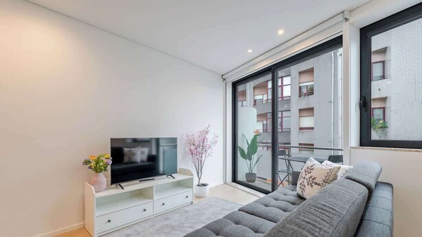 Una sala de estar moderna con un sofá gris, un televisor de pantalla plana sobre un soporte blanco, una planta en maceta y un ventanal con vistas a un edificio.