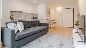 Una sala de estar moderna con un sofá gris, una pequeña mesa de comedor y una zona de cocina con armarios blancos y un microondas.