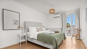 Een slaapkamer met een bed, nachtkastje, lamp en uitzicht op de omgeving door een groot raam.