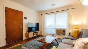 Una sala de estar con puerta de madera, televisor, sofá, mesa de centro y ventana con persianas.