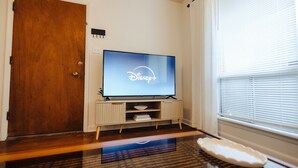 Una sala de estar con una puerta de madera, un televisor mostrando el logotipo de Disney+, un mueble blanco y una ventana con persianas.