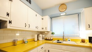 Una cocina con armarios blancos, encimera amarilla, un lavamanos, un reloj y una ventana con persianas.
