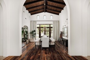 Un comedor con piso de madera, paredes blancas y un techo alto con vigas a la vista. Se observa un ventanal con cortinas, una araña y una mesa de comedor con sillas.