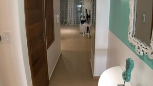 Un couloir avec une porte en bois, un mur blanc orné d’un miroir décoratif et un lavabo blanc surmonté d’une sculpture turquoise.