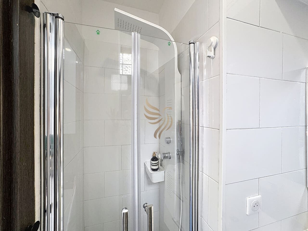 A glass-enclosed shower with a white tiled wall, a chrome shower head, and a small shelf with toiletries.