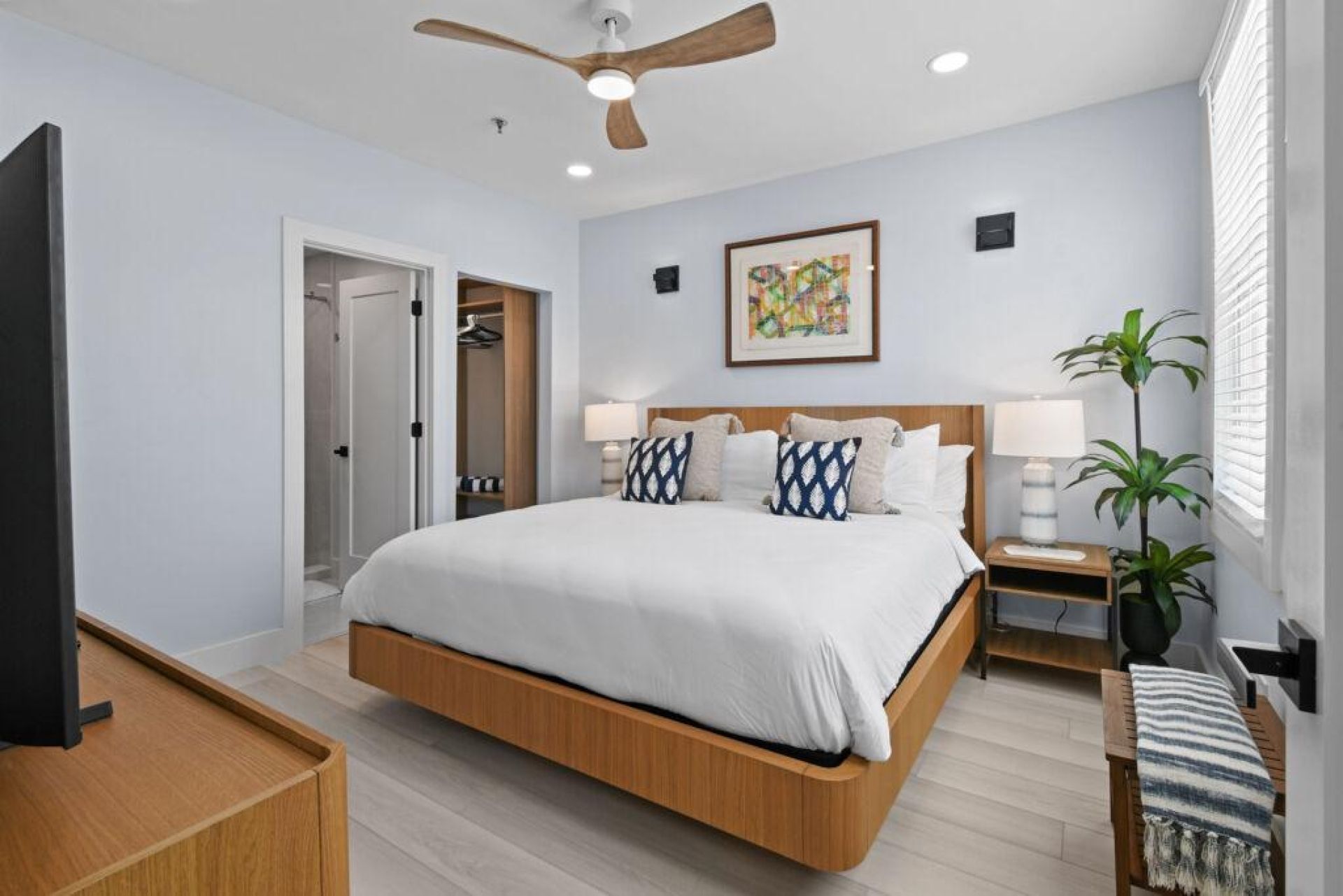 Une chambre d’hôtel avec un lit, des tables de chevet, un ventilateur de plafond, un tableau et une plante en pot.