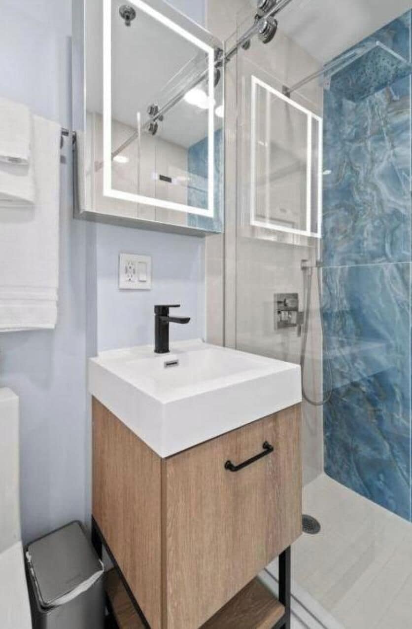 Une salle de bain moderne avec une douche à l’italienne en verre, un lavabo blanc doté d’un robinet noir et un pare-baignoire en marbre.
